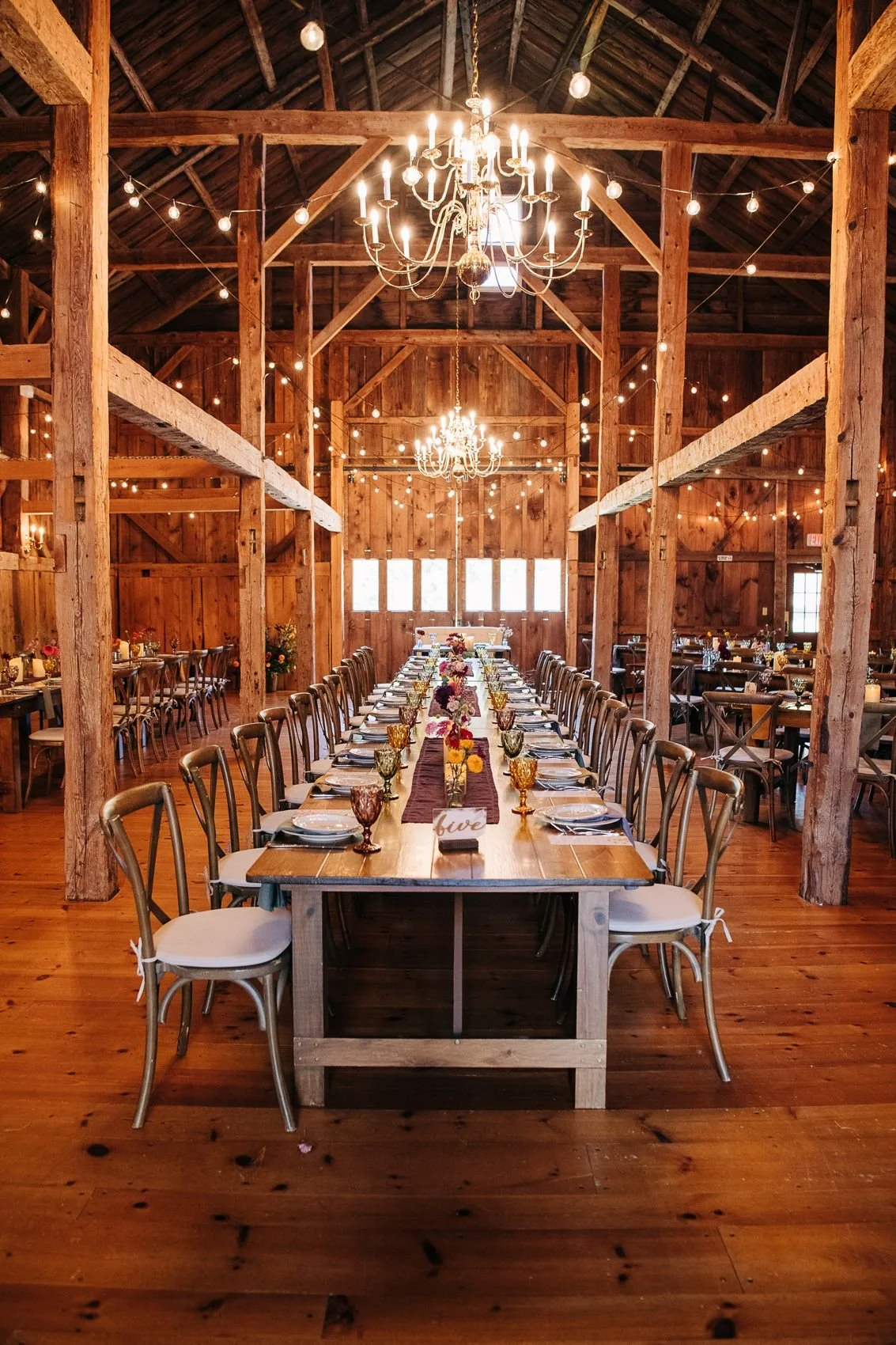 Long reception tables set beneath chandeliers and glowing string lights inside the timber-frame barn at William Allen Farm