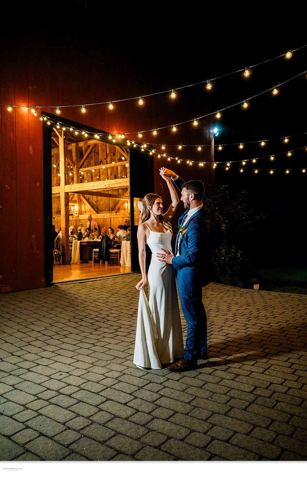 A couple dancing outside the glowing barn under string lights at night