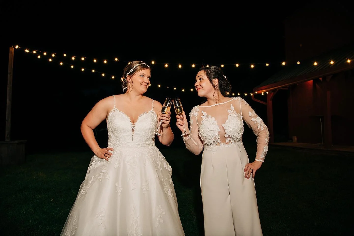 Two brides smiling as they toast with champagne under string lights beside the barn at night