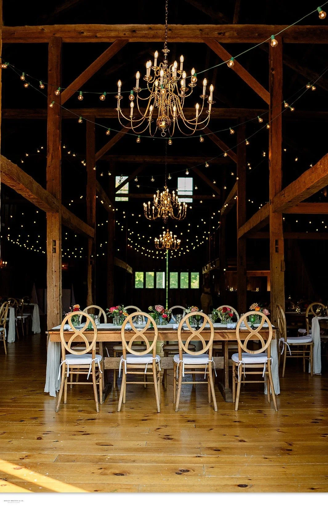 Set farm tables beneath chandeliers and a canopy of string lights in the barn