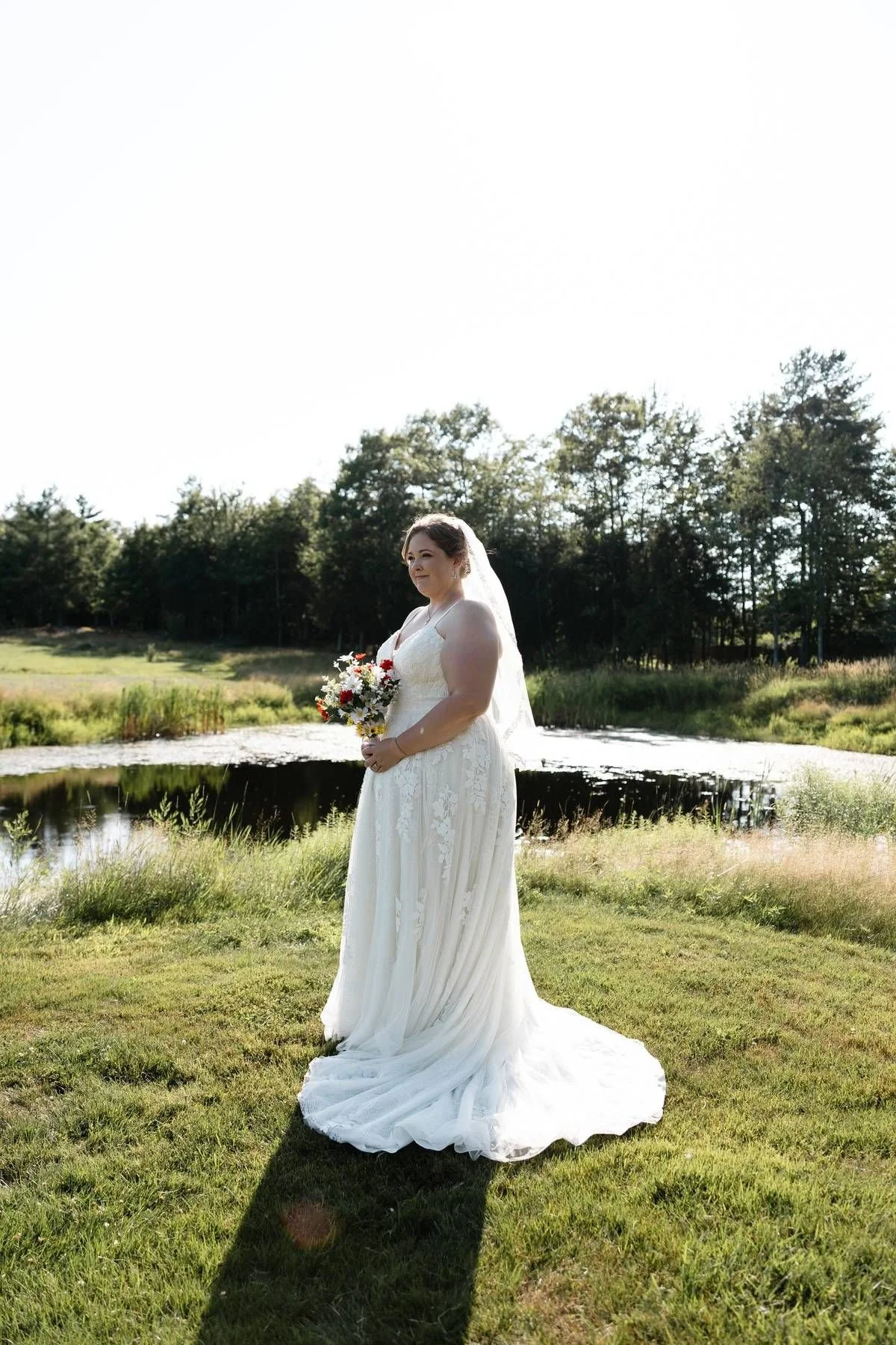 A bride in a long veil standing beside the pond at William Allen Farm