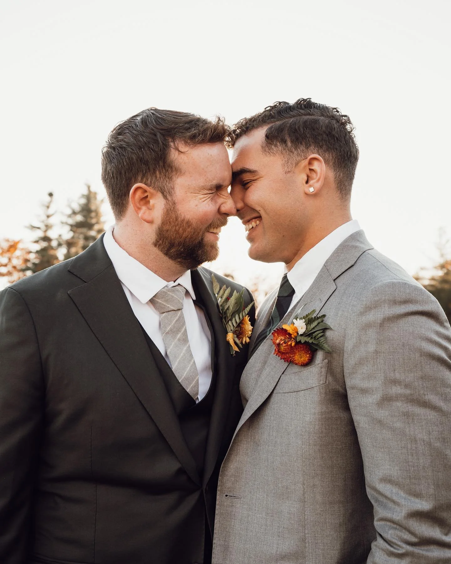 Two grooms touching foreheads and smiling among the pines at William Allen Farm