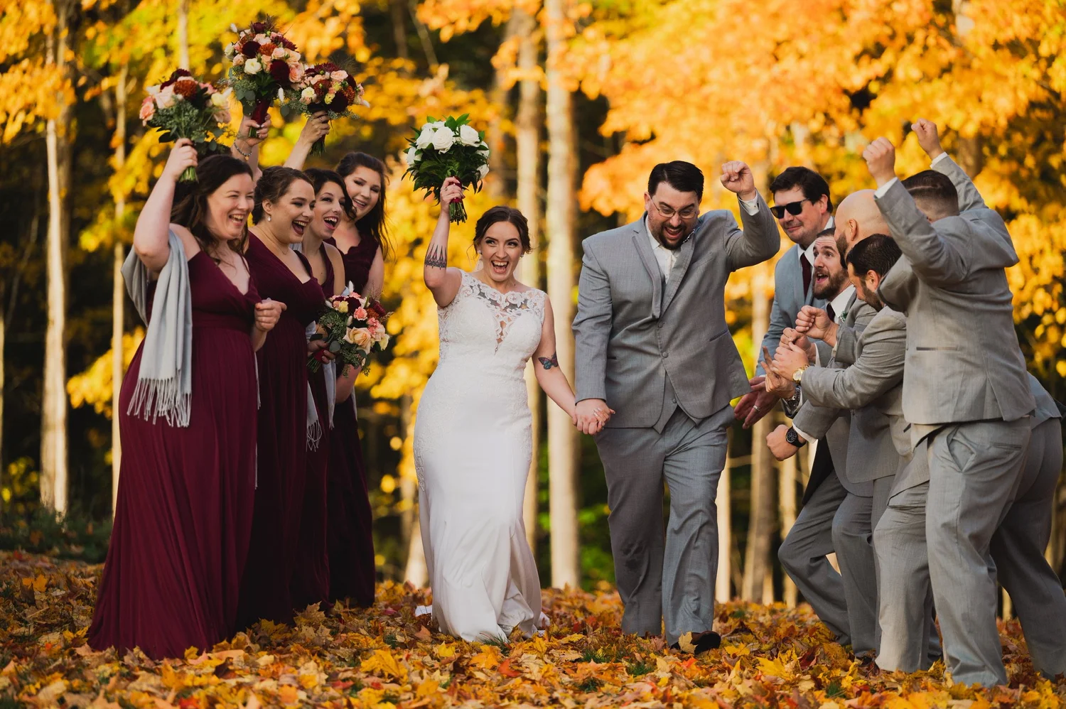 A bride and groom walk hand in hand through fall leaves as their cheering wedding party raises bouquets at William Allen Farm