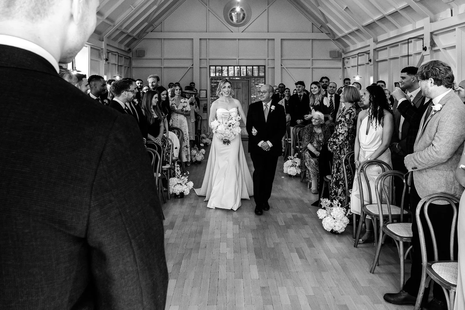 Abi walking down the aisle with her father at Limekiln, guests watching on both sides, flowers lining the aisle — black and white