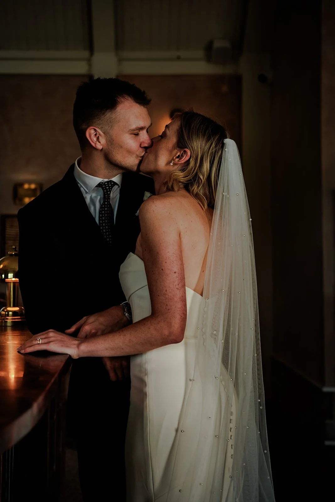 Jack and Abi sharing a quiet, intimate kiss at the bar in warm candlelight at Limekiln — atmospheric and romantic evening portrait