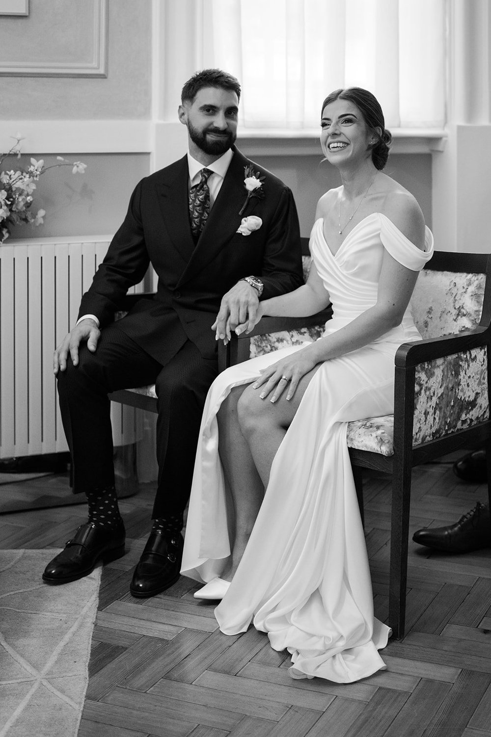 Bride and groom sitting together on a wooden bench beside a tall window at Chelsea Old Town Hall