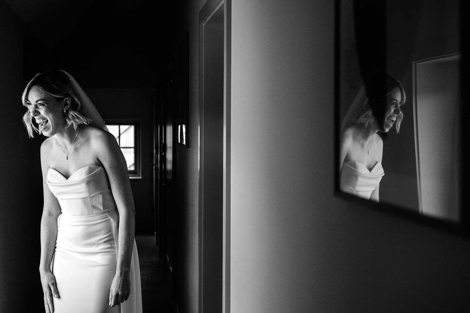 Bride Abi in her strapless white wedding dress laughing with pure joy, reflected in a dark-framed mirror at Limekiln — striking black and white portrait
