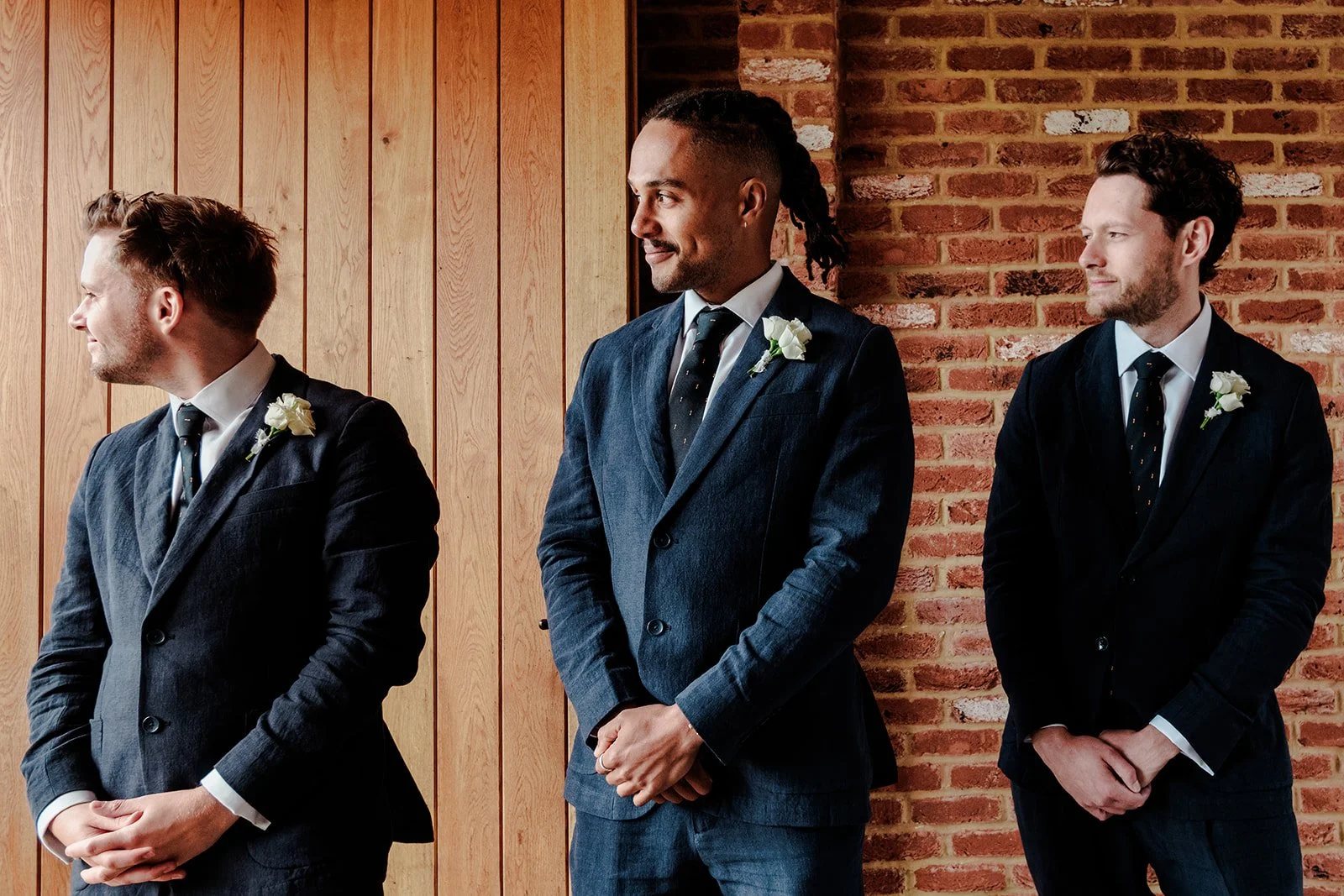Groom Jack and his groomsmen in dark suits waiting for the ceremony to begin, standing against the brick wall inside Limekiln's ceremony barn