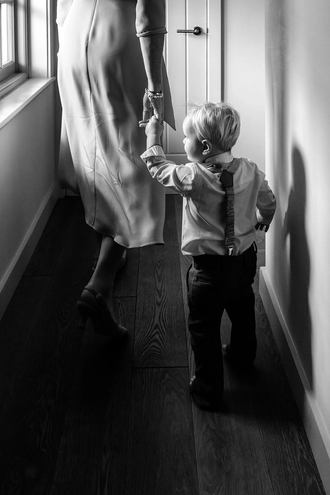 Adorable young pageboy in a smart suit and braces holding a grown-up's hand in the hallway — perfectly timed black and white wedding moment