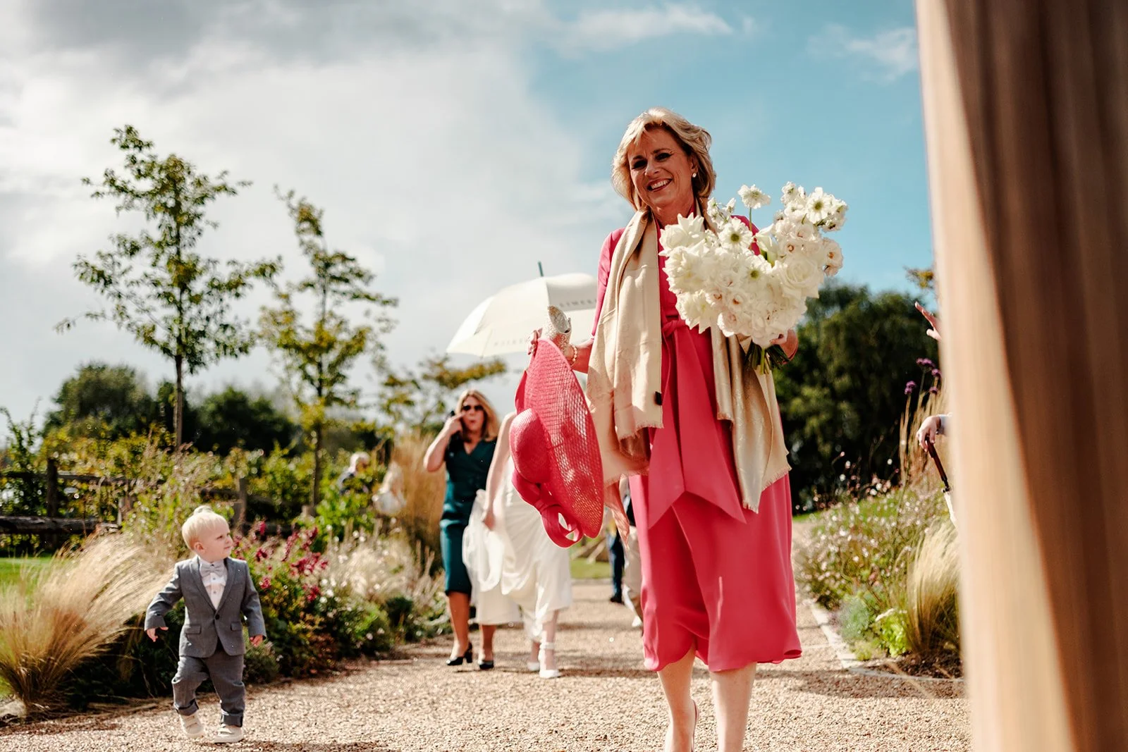 Outdoor wedding procession at Limekiln — elegantly dressed guest in coral-pink carrying a bouquet of white flowers, with the young pageboy running alongside