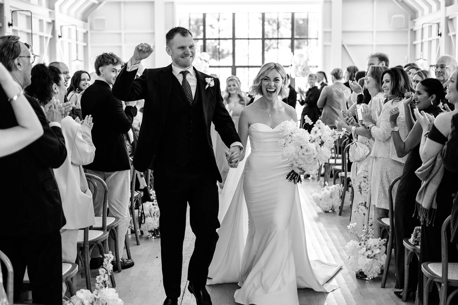 Newly married Jack and Abi walking triumphantly back down the aisle, arm in arm, while guests cheer on both sides — black and white