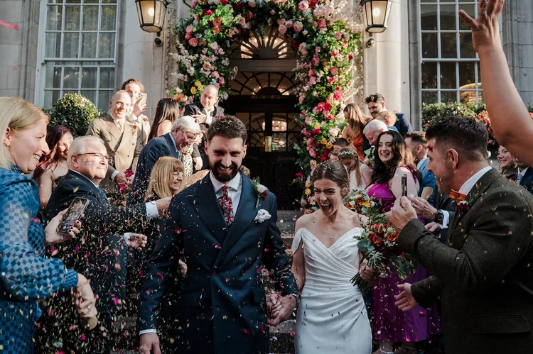 Nat and Josh under the flower archway at Chelsea Old Town Hall in a shower of confetti