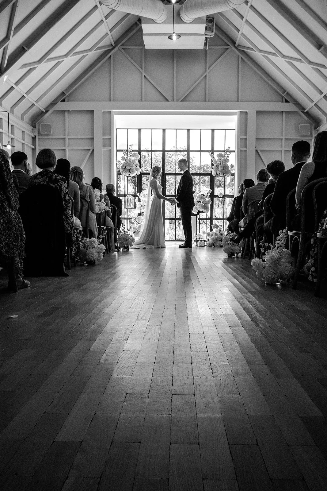 Wide-angle black and white shot of Jack and Abi's ceremony at Limekiln — the couple silhouetted against the tall arched windows, guests lining both sides of the barn