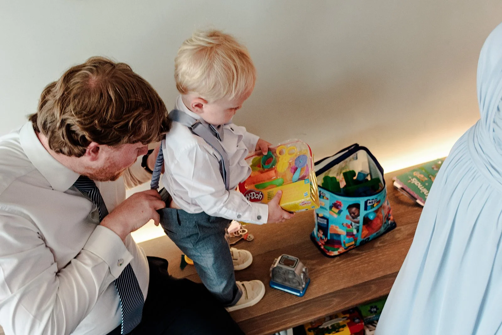 Groom crouching down to help the little pageboy with his outfit while toys are spread across the table — candid getting-ready moment at Limekiln