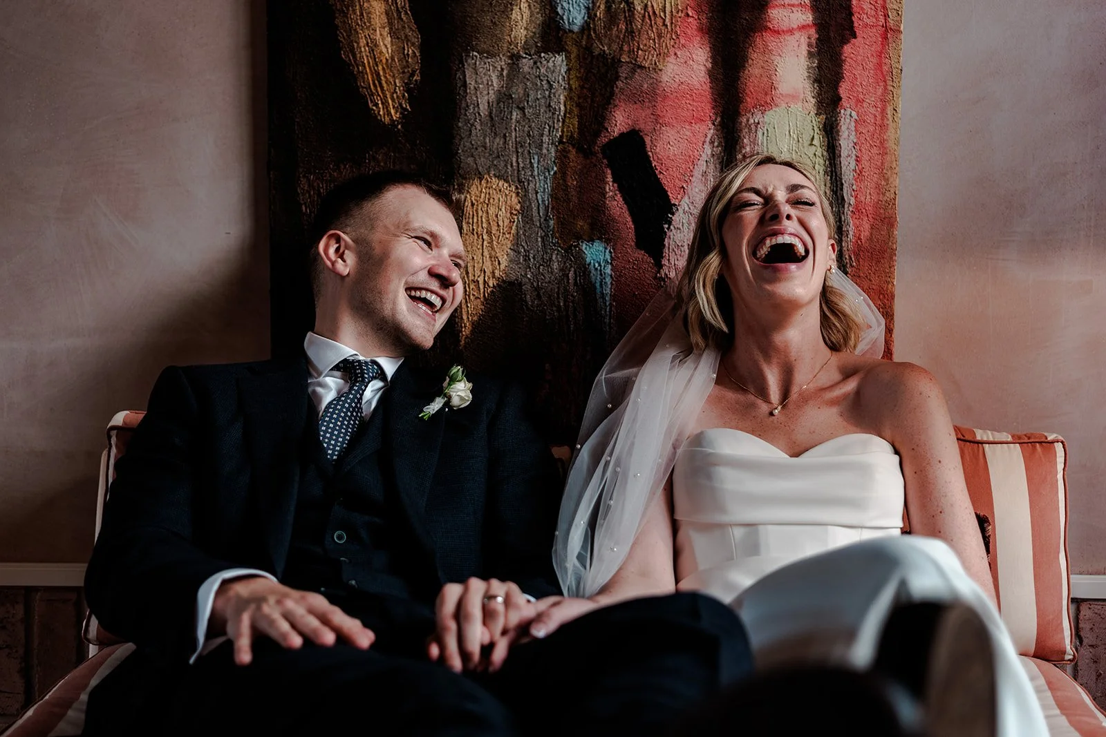 Jack and Abi laughing together on a velvet sofa in front of a colourful abstract artwork inside Limekiln — warm, relaxed couple portrait
