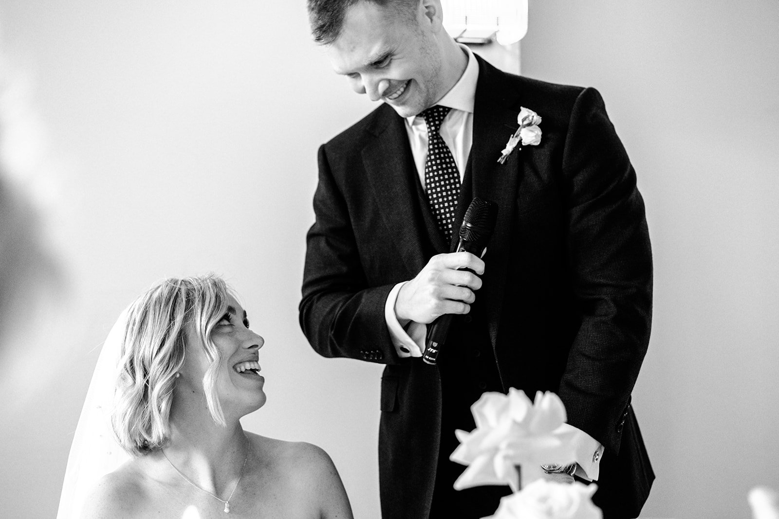 Groom Jack delivering his wedding speech at Limekiln, microphone in hand, while Abi looks up at him laughing — beautiful candid moment, black and white