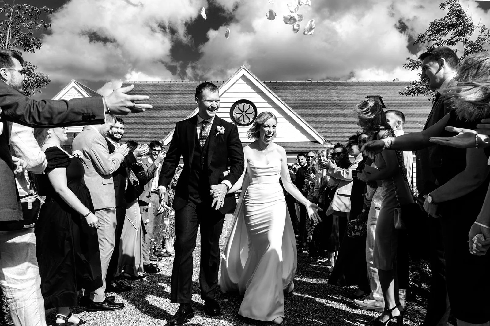 Jack and Abi walking through a confetti shower outside the Limekiln ceremony barn, guests throwing confetti under a dramatic cloudy sky — black and white