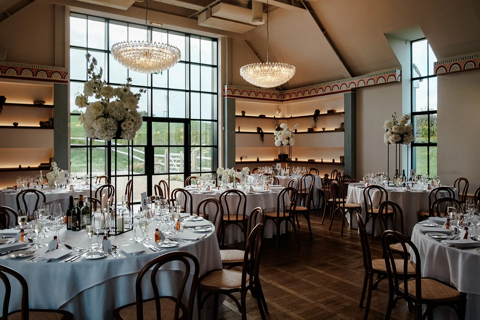 The stunning Limekiln dining room set for the wedding breakfast — crystal chandeliers, tall windows, round tables with white linen and tall white floral centrepieces