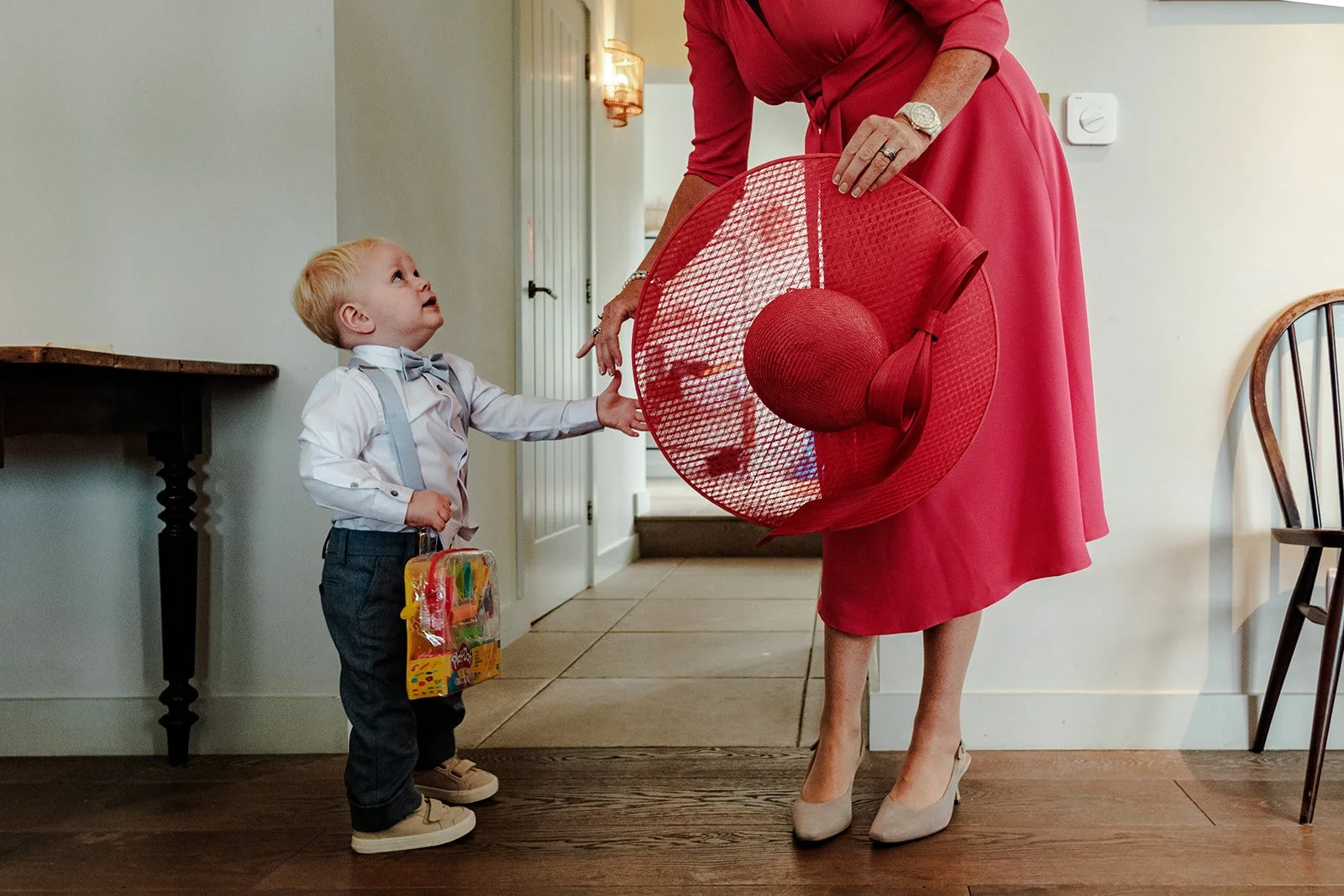 Small boy in a wedding suit reaching up to inspect a woman's large red hat in the hallway at Limekiln before the ceremony