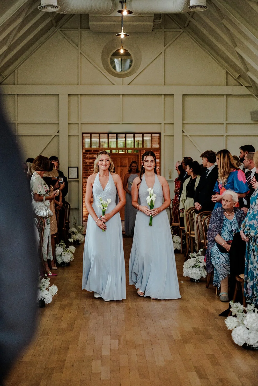 Two bridesmaids in pale blue halter-neck dresses walking down the aisle together inside Limekiln's beautiful ceremony barn