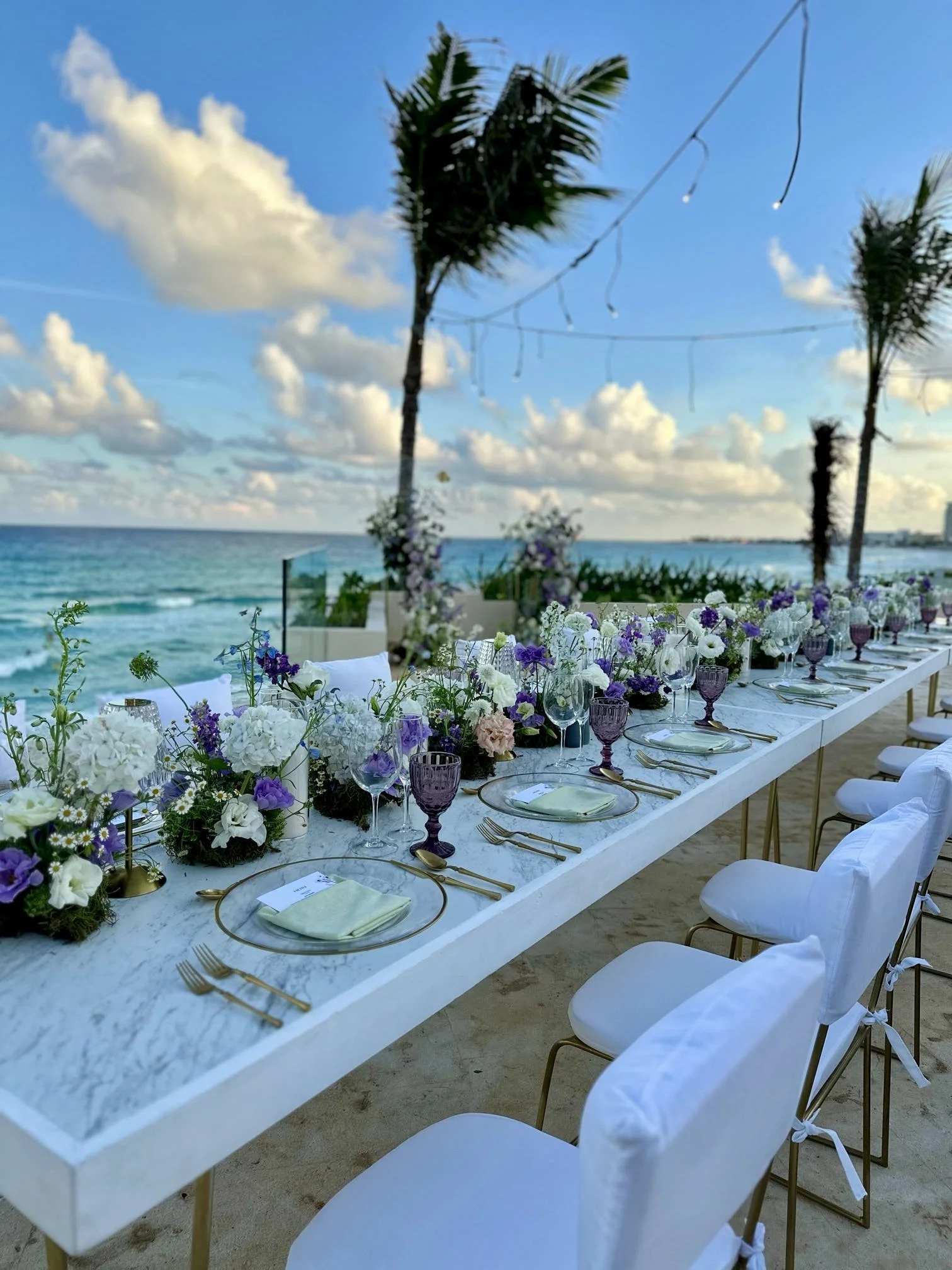 Long beachfront wedding reception table set with flowers, glassware, and purple accents by the ocean