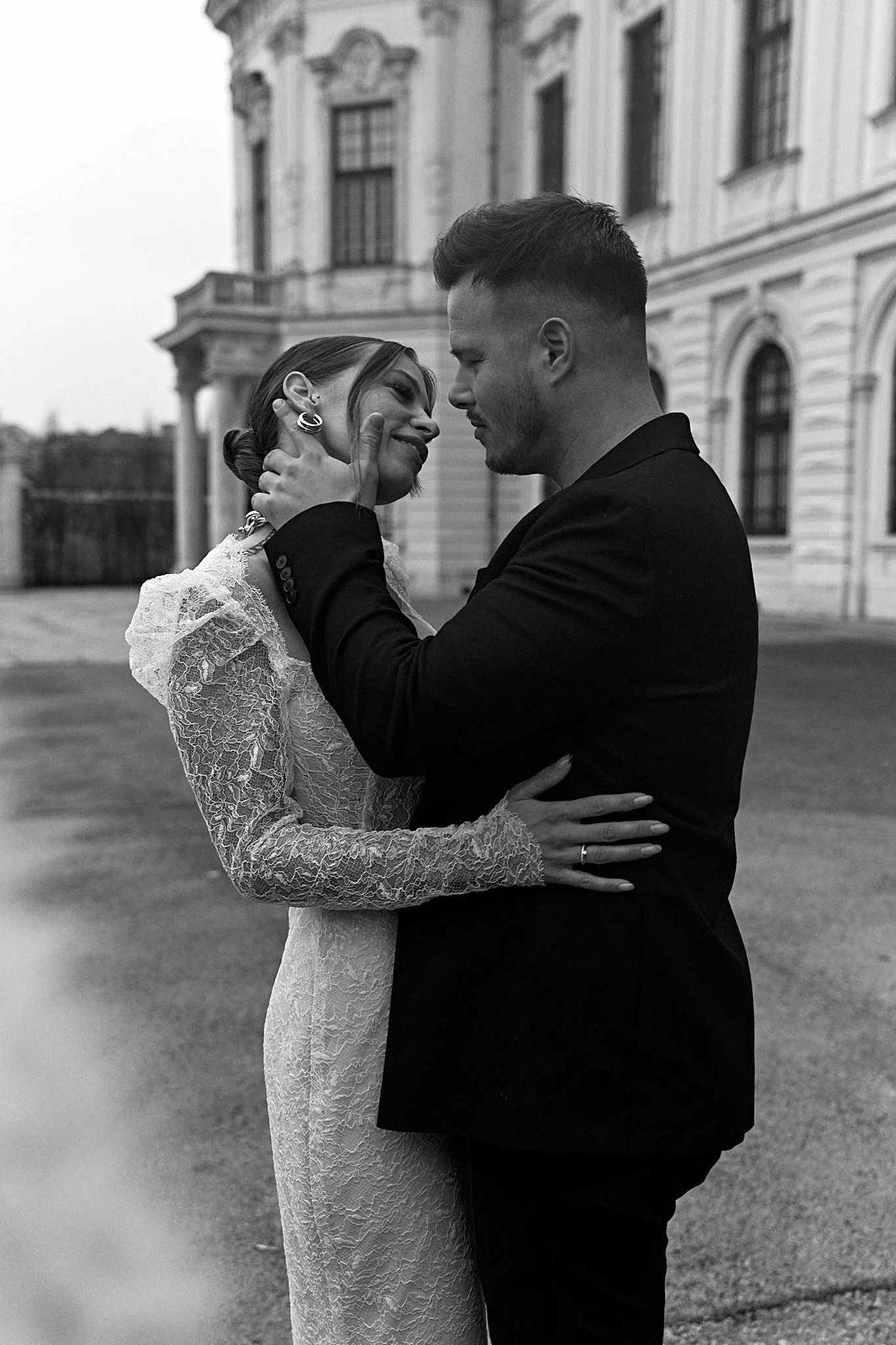 A bride in a lace dress smiling as her partner holds her face outside a palace, photographed by Love Society Studios