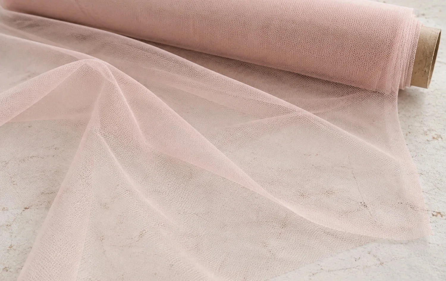 Plain blush-pink illusion tulle draped from a fabric roll, showing its fine open structure.