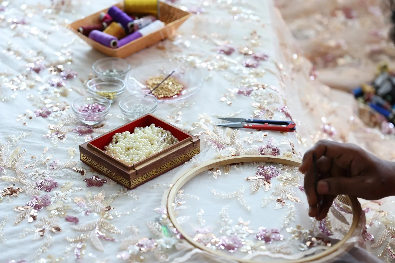 An artisan stitching floral embellishment in an embroidery hoop, beside trays of beads, sequins and coloured thread.