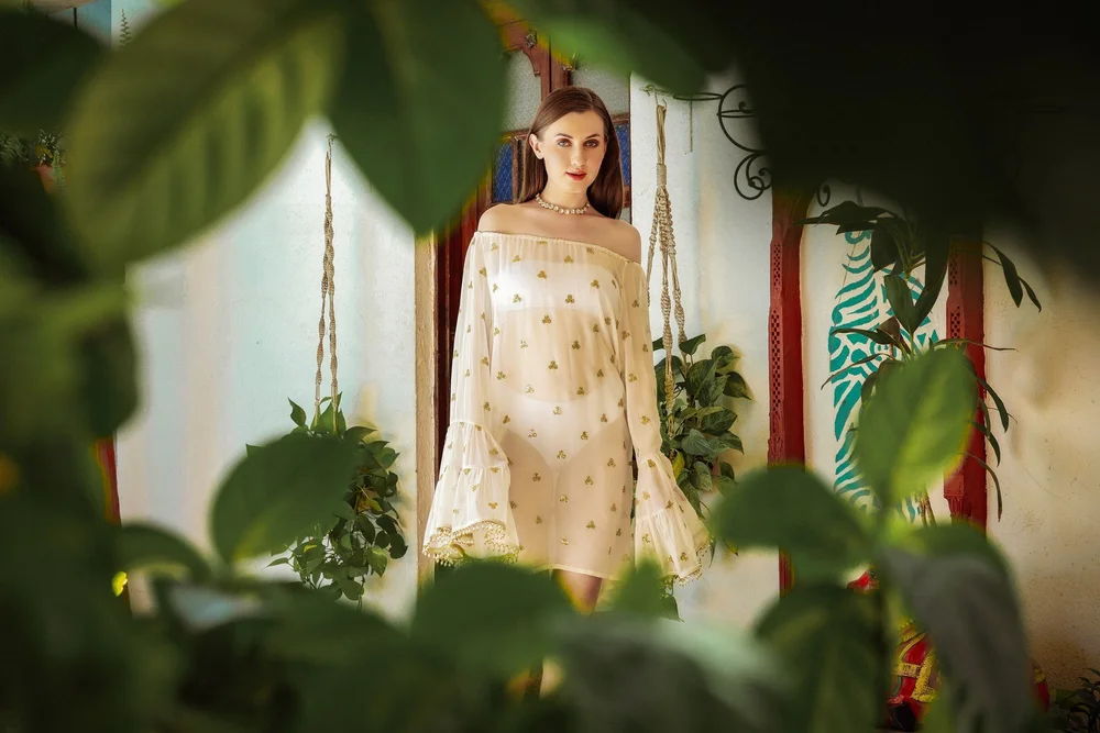 Pearl Maharani Dress photographed through foliage, showing the off-shoulder neckline and gold embroidery.