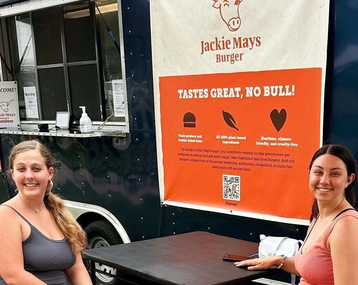 Two smiling customers at the Jackie Mays Burger food truck, with the "Tastes Great, No Bull!" banner in the background