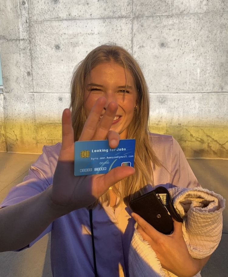 Kyra smiling in golden-hour light, holding up a blue card that reads Looking for Jobs