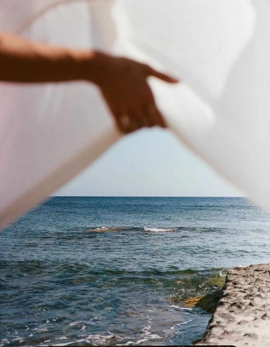 A hand lifts a sheer curtain to reveal a calm ocean