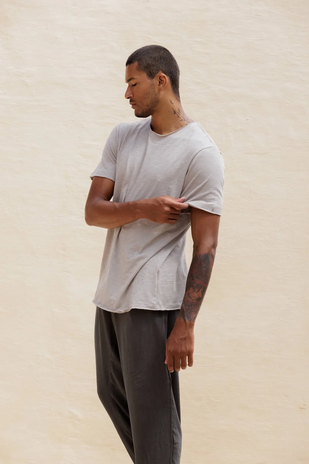Man wearing Nousa Concept SS26 grey cotton t-shirt and dark trousers, sand wall background