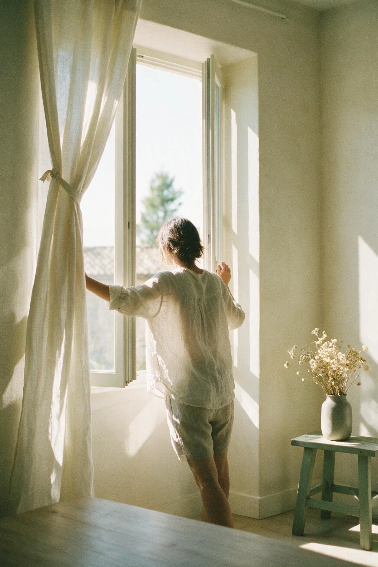 Une femme de dos ouvre une fenêtre, le regard vers l'extérieur lumineux.