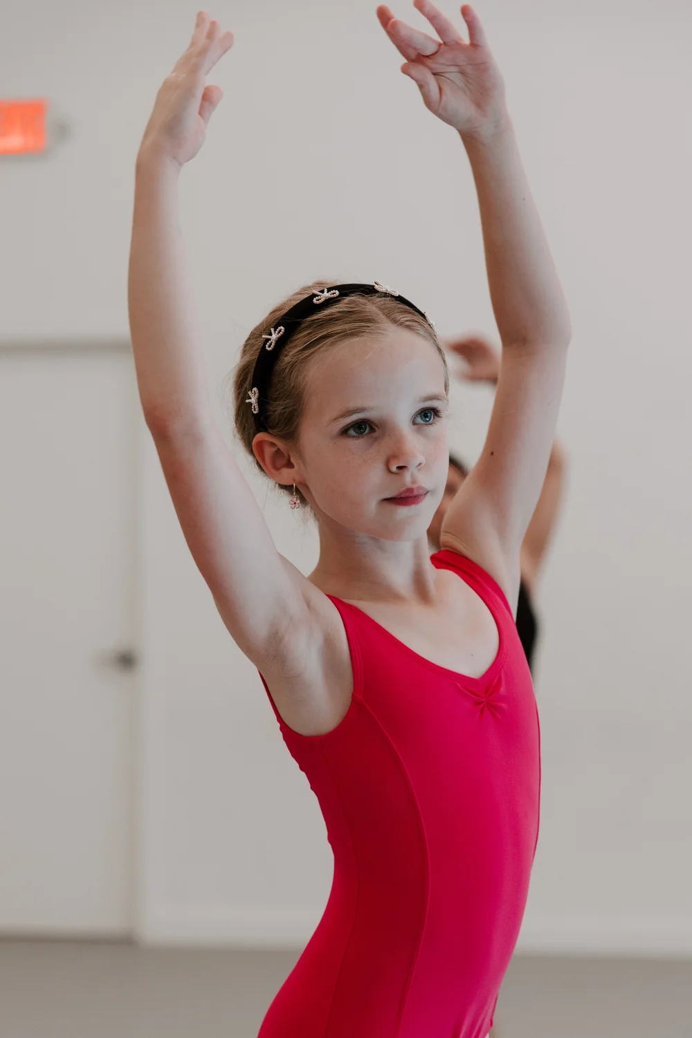 A young dancer in a red leotard holding her arms in fifth position during class