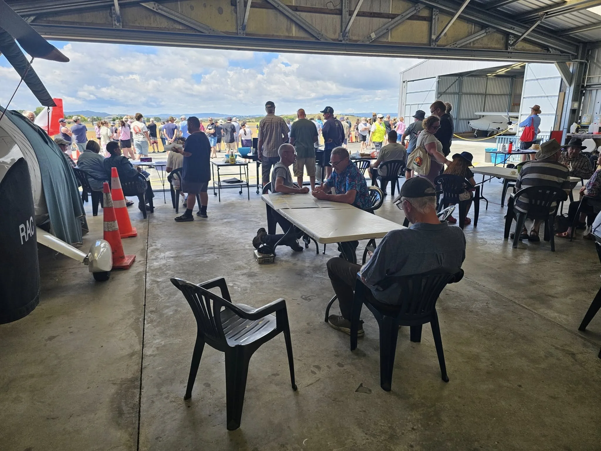 Whangarei Flying Club members socialising inside a hangar during a public aviation event in Northland, New Zealand, with aircraft and crowd visible in the background.