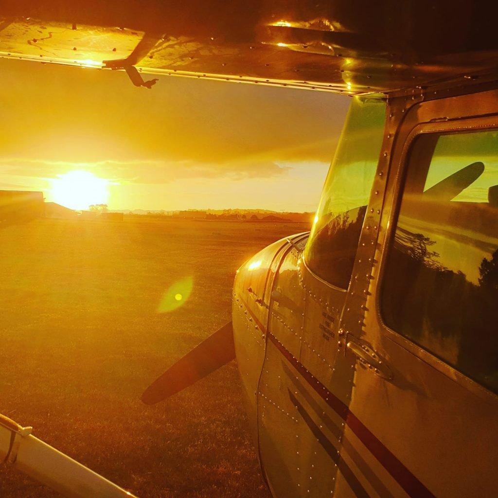 Small aircraft parked on the runway at sunrise, with the warm orange glow illuminating the New Zealand landscape in the background.