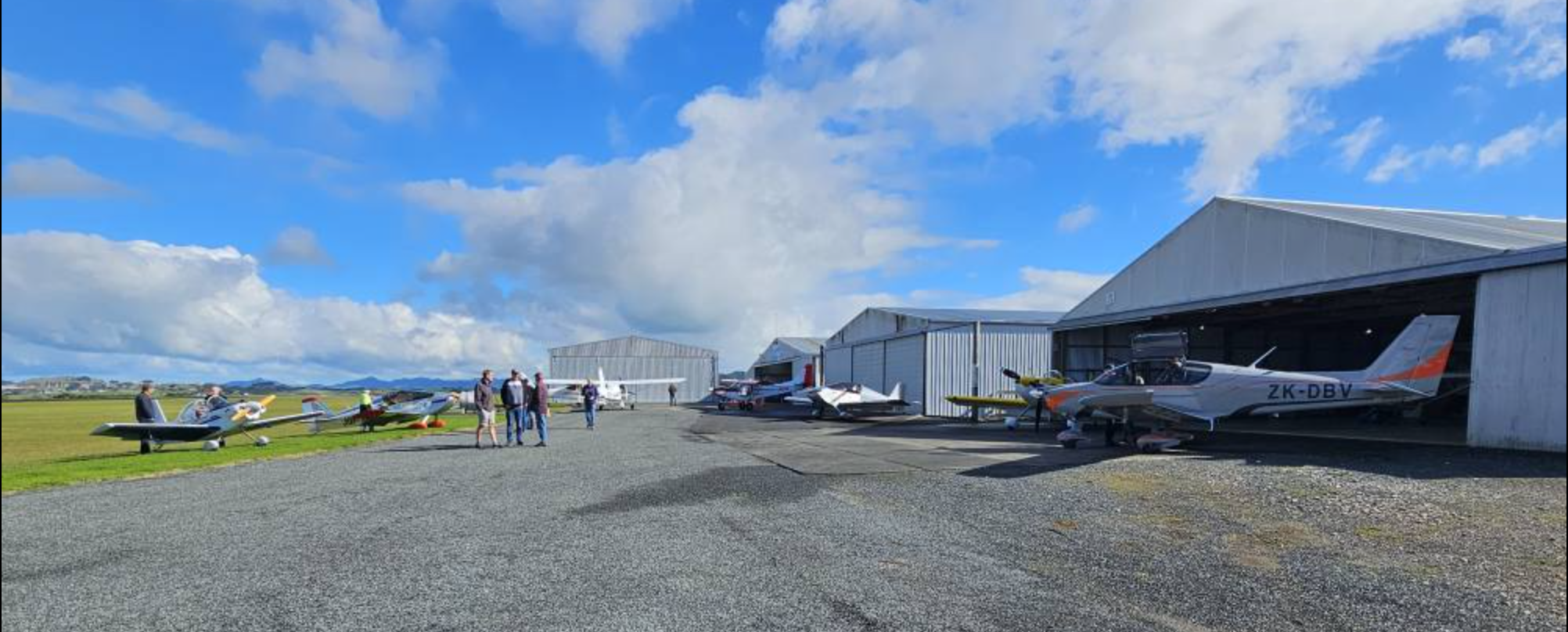 Light aircraft parked outside hangars at Whangarei Flying Club in Northland, New Zealand, with club members interacting under a bright blue sky.