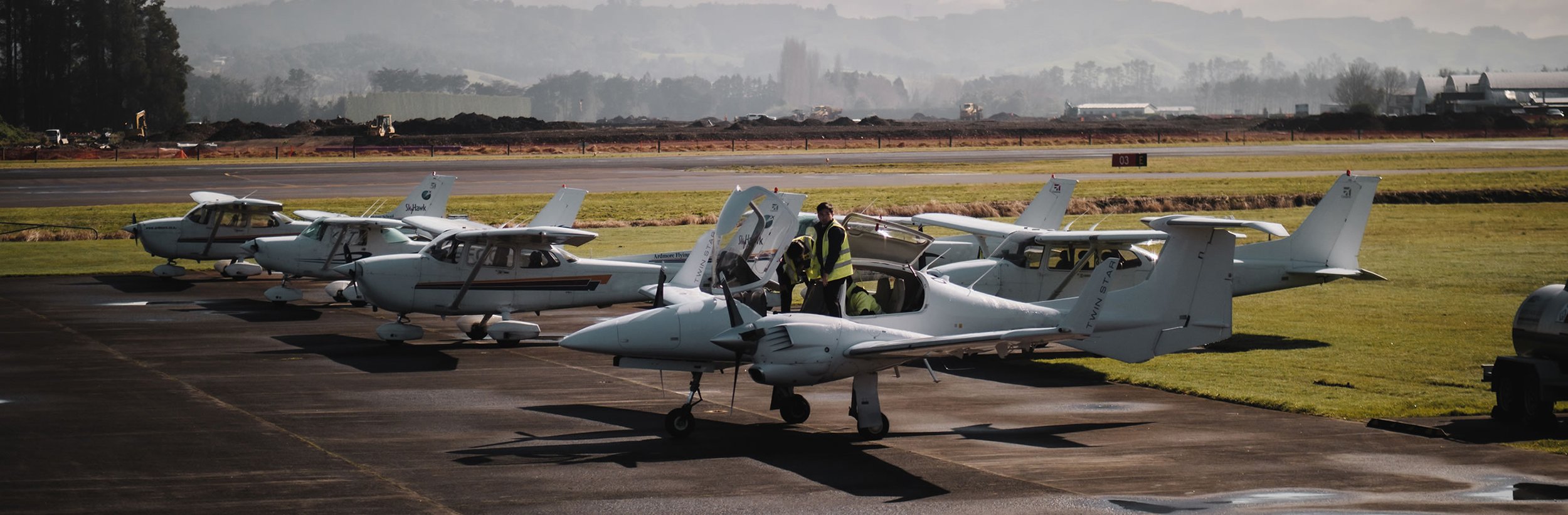 Three trainee pilots in high-visibility vests walking past a Cessna Skyhawk on the Ardmore Flying School apron in Auckland.