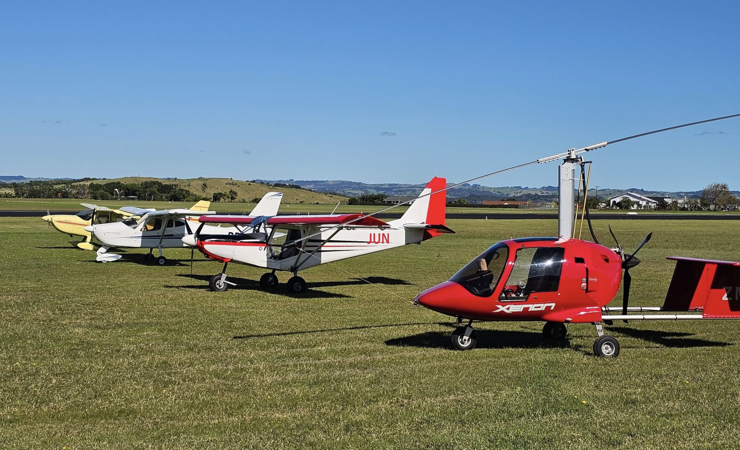 Savannah S aircraft ZK-JUN and other light sport aircraft parked on the grass at Whangarei Flying Club in Northland, New Zealand, on a clear day.