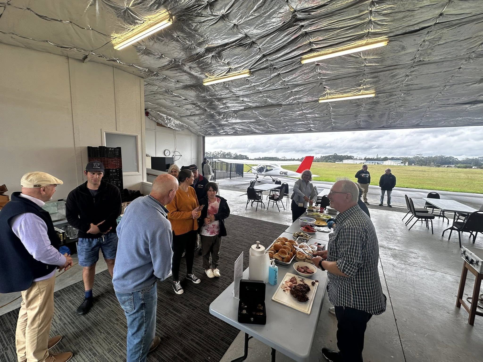 Bay of Islands Aero Club members enjoying food and conversation inside a hangar at Kerikeri Airport, with a light aircraft and airfield visible in the background.