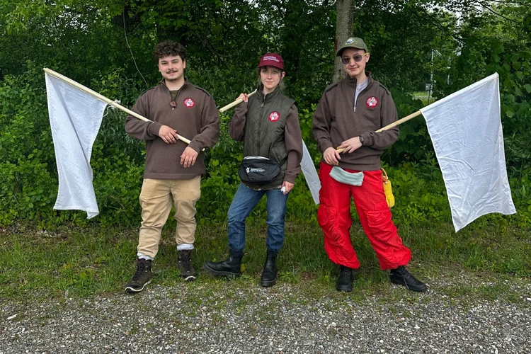 Three people hold white flags and pose side-by-side wearing NEWVEC-branded quarter-zips. 

Dr. Rich gives a presentation to Americorps members at a projector screen in a mostly empty room.

A volunteer bends down to carefully inspect a flag for tick.