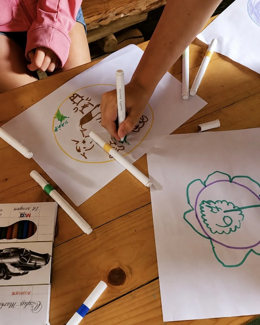 Children's hands drawing with markers on large sheets of paper on a wooden floor.