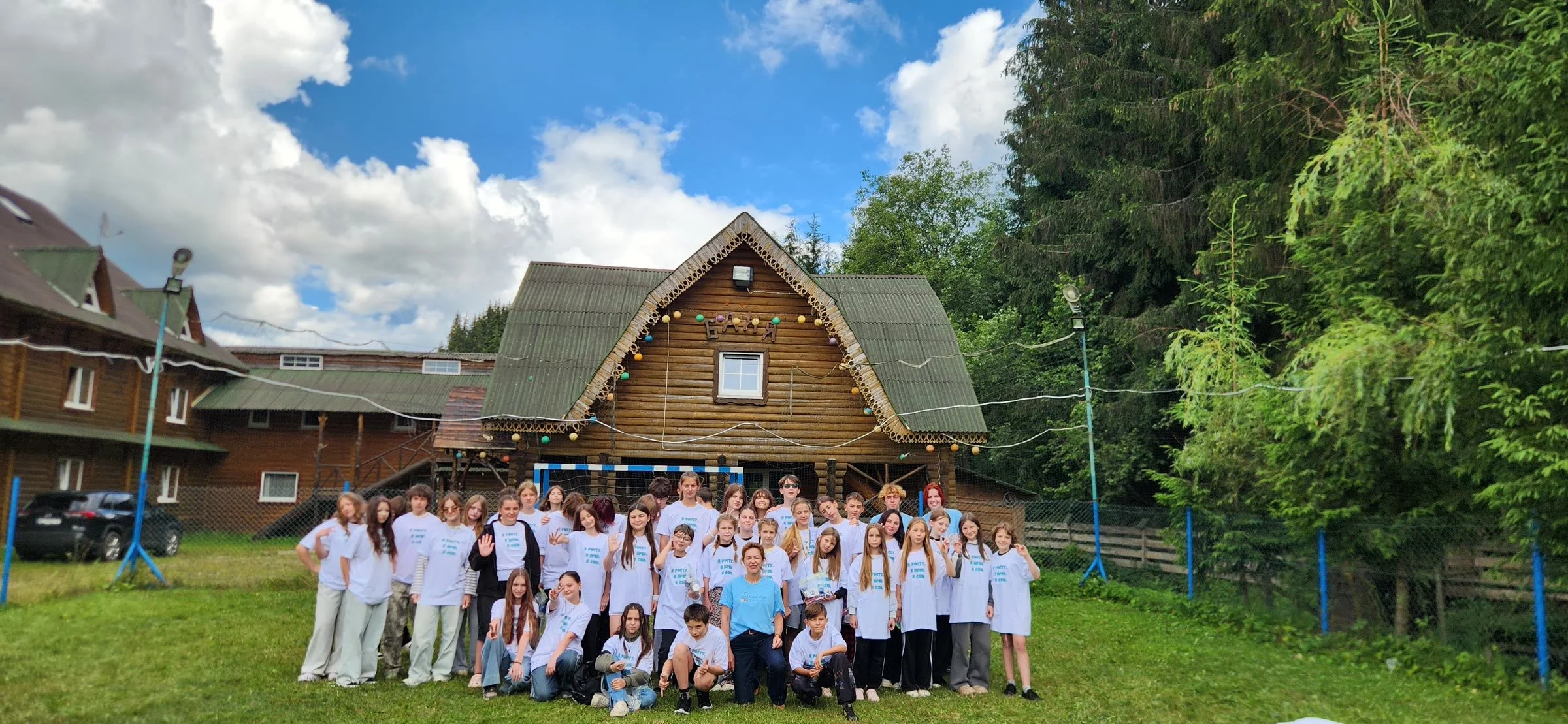 About fifty children and young volunteers in matching white T-shirts stand together on the grass in front of a wooden mountain lodge, under a blue sky with white clouds.