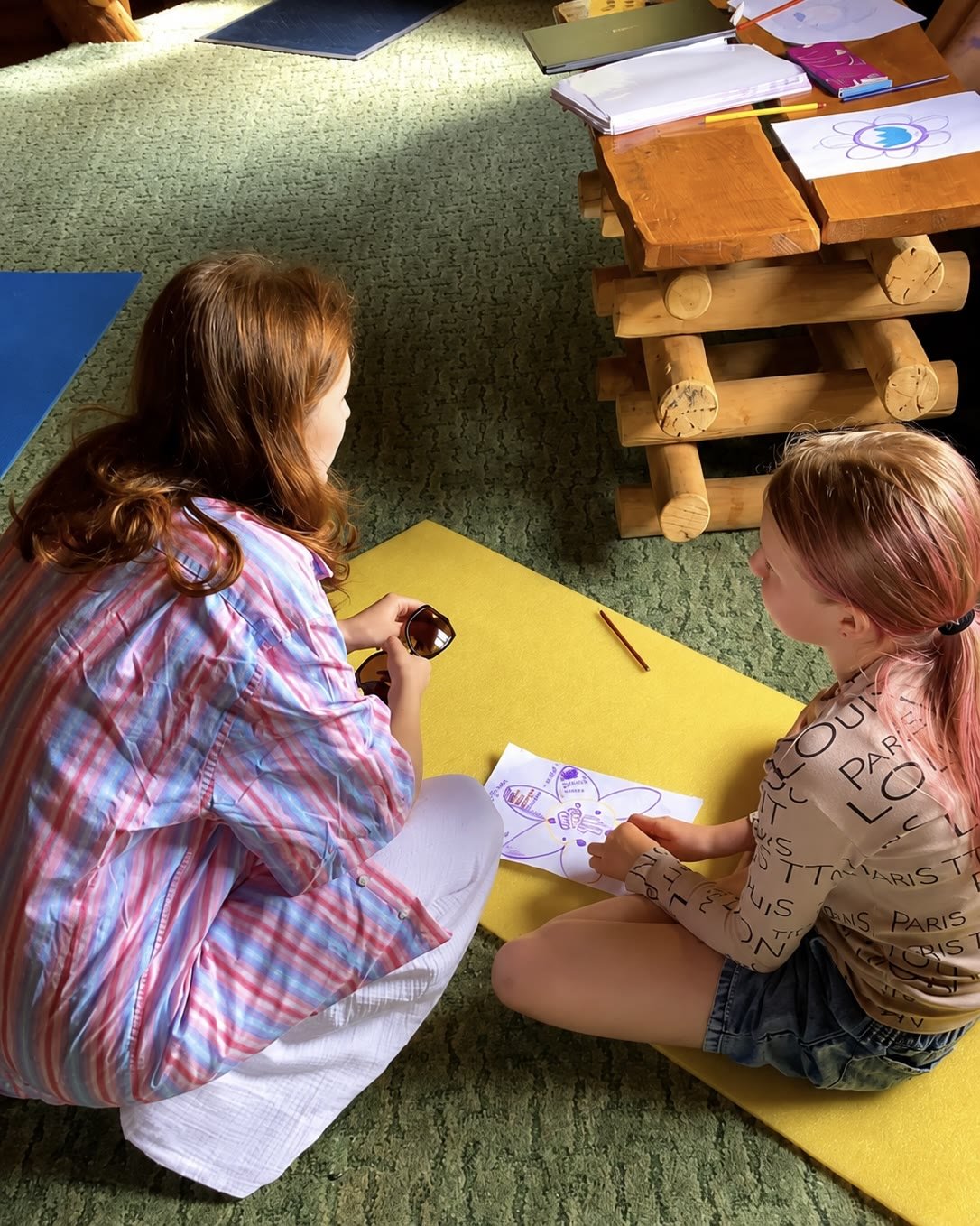 A young woman sits cross-legged on a yellow mat beside a girl, looking together at a drawing.