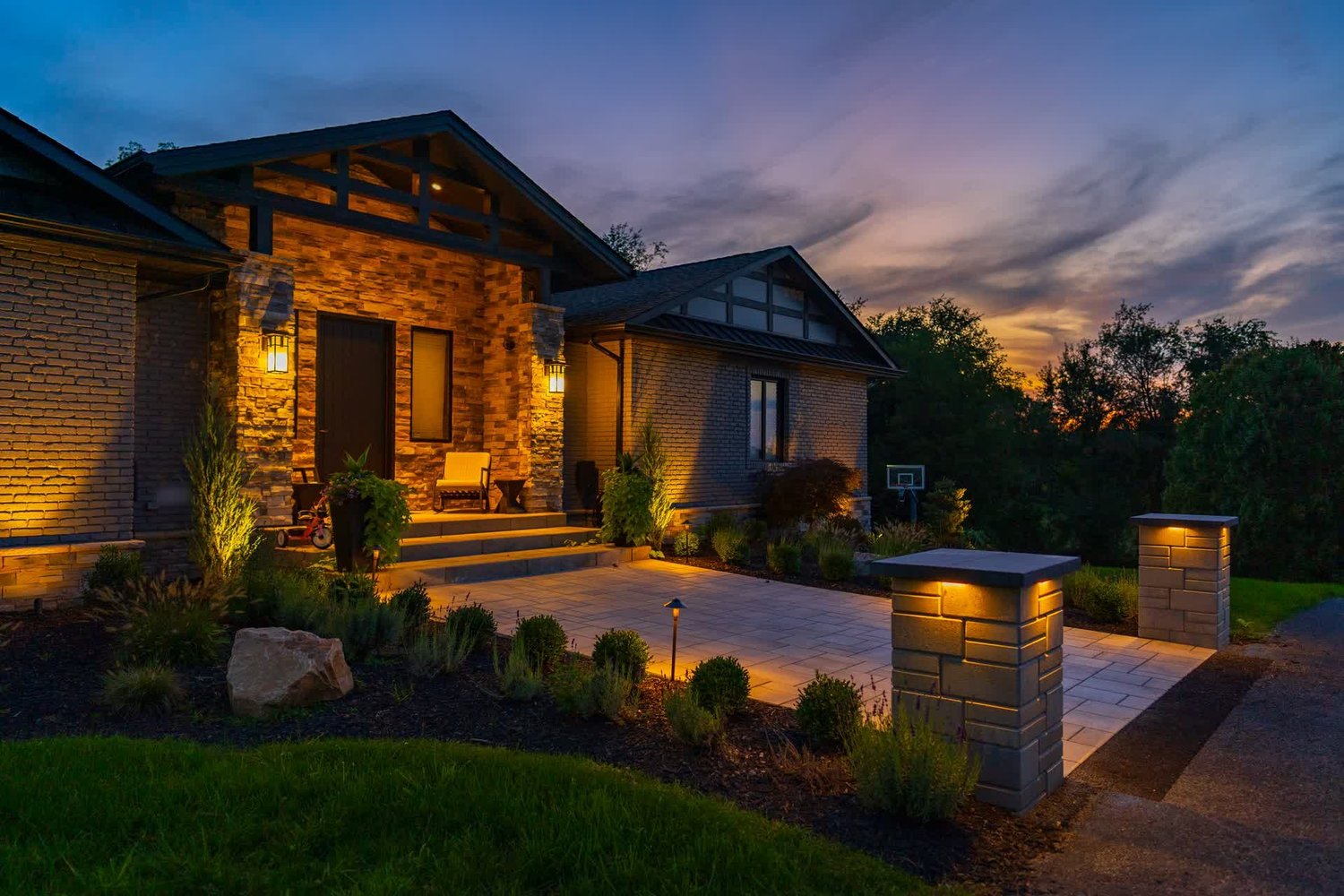A lit stone walk and pillars leading to a front entry at dusk
