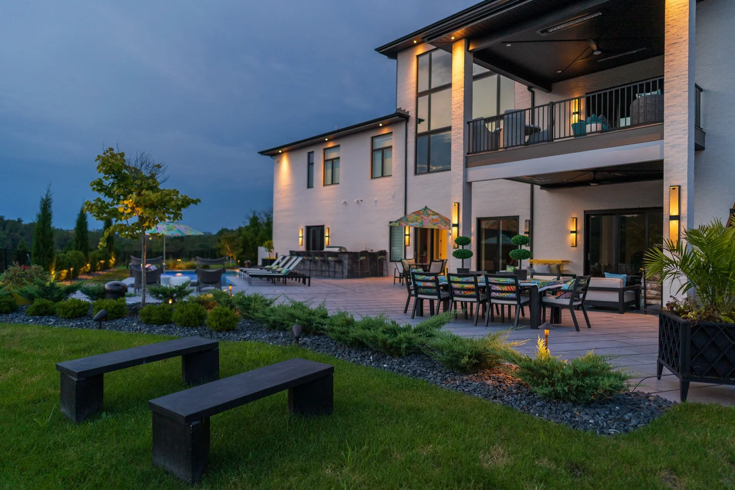 The back of the house, the patio and the dining area at dusk, with benches on the lawn
