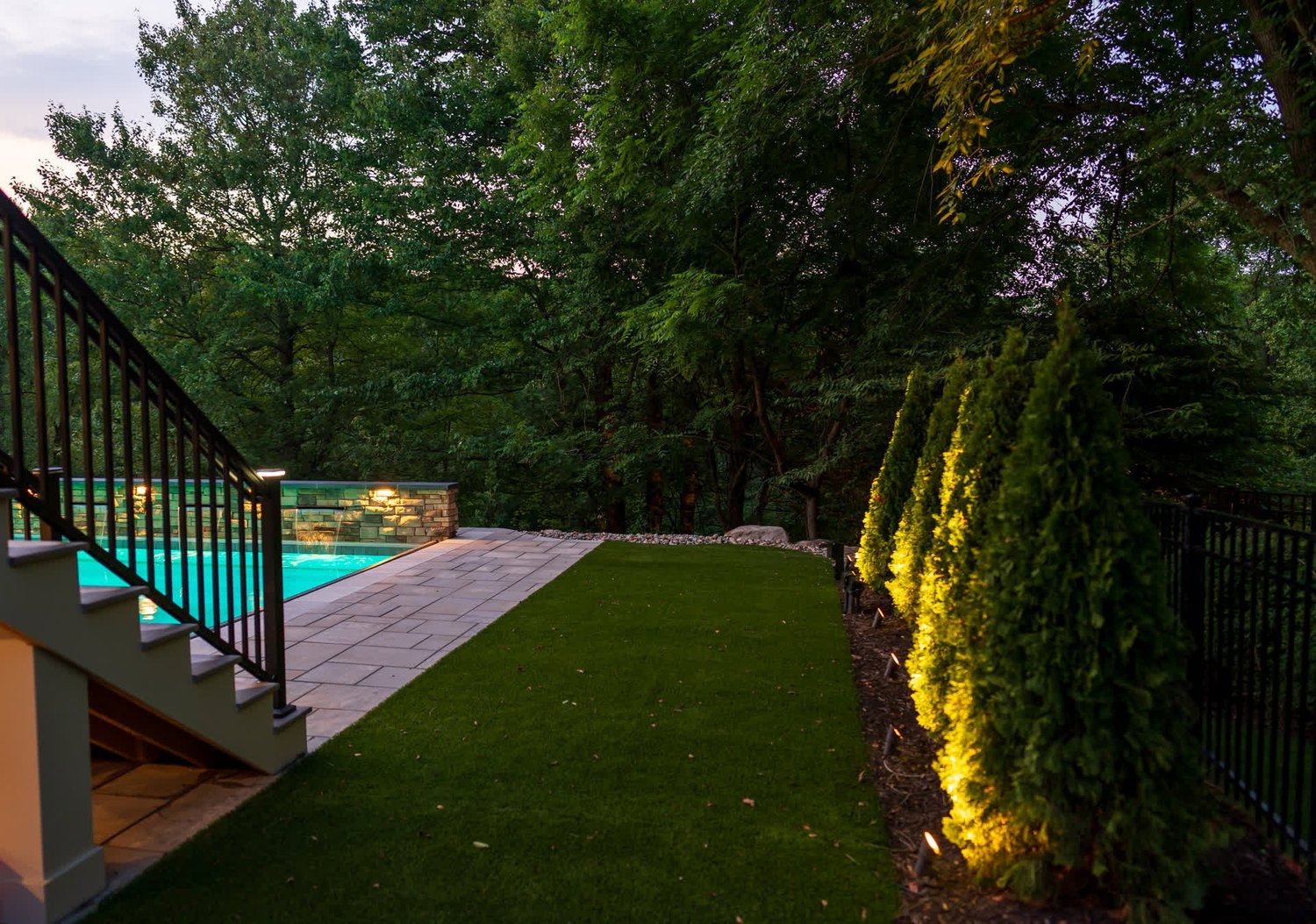 Steps beside a lit row of evergreens, down to a lawn and a pool