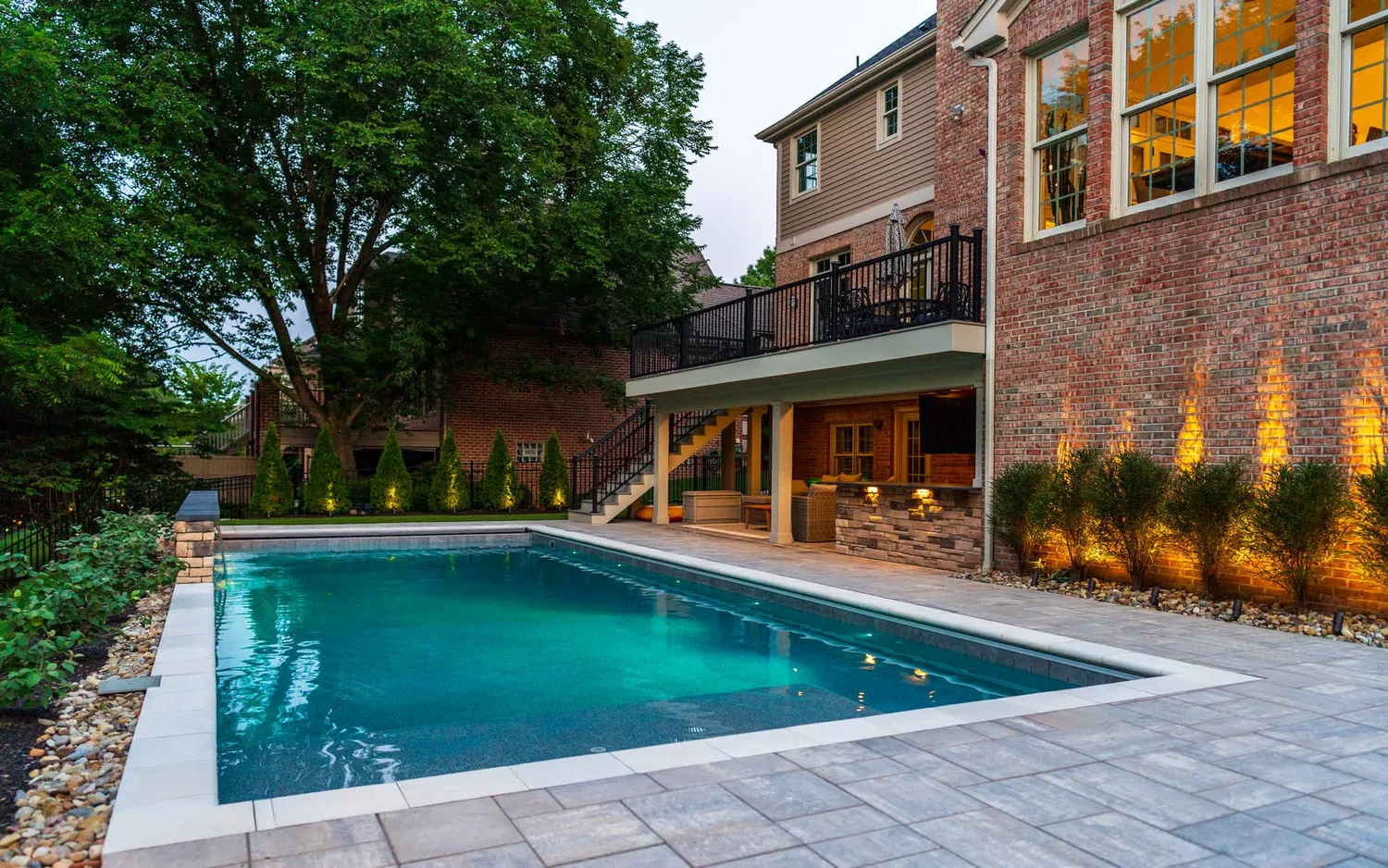 A pool beside a brick house, lit plantings along the wall