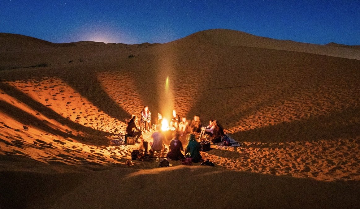 Group of people gathered around a fire in a sand dune at night
